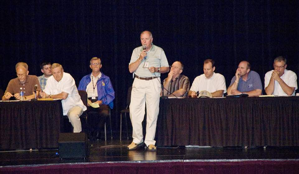 Residents seated in rows at a town hall meeting, listening to a speaker at the front of the room.